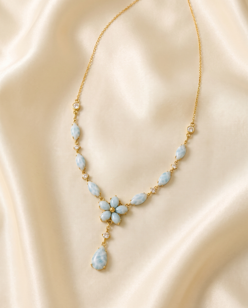 A gold chain necklace with light blue marbled stone accents and a teardrop pendant, displayed on draped cream satin fabric.